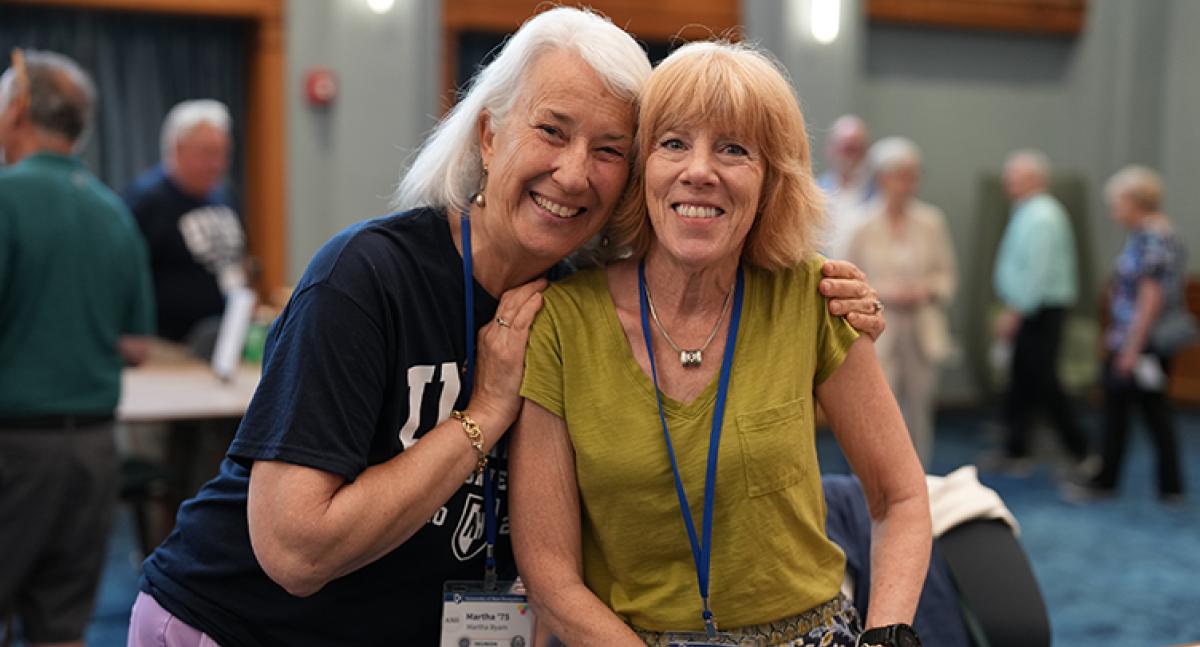 A woman puts her hands on the shoulders of another woman and leans in close for a photo