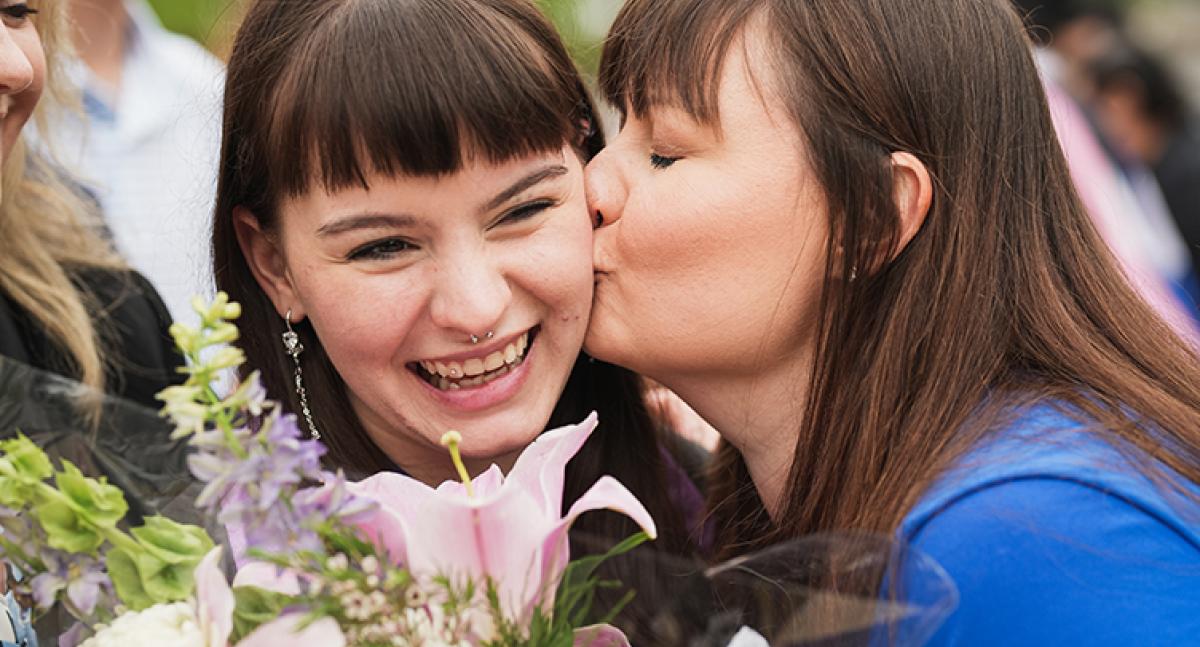 A graduate holding flowers smiles while getting a kiss on the cheek from another woman