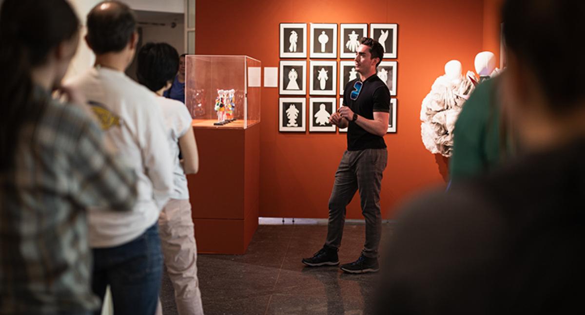 A man in front of an orange wall speaks to a small standing crowd