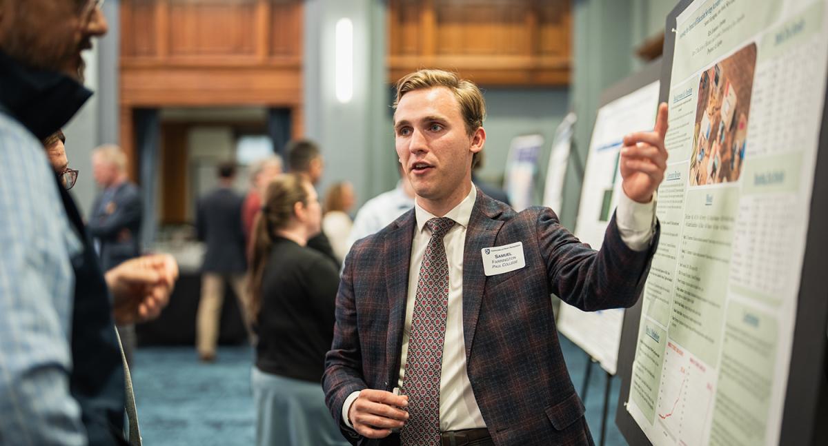 A man in a suit and tie points toward a poster while speaking to another man