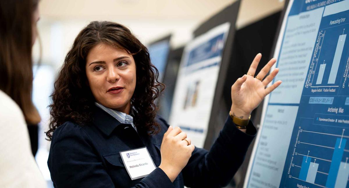 A woman looks back while speaking to a guest as she explains research on a poster in front of her