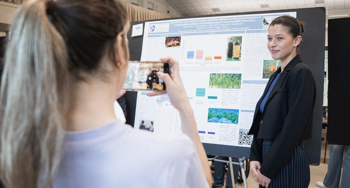 A woman with a ponytail takes a picture on her cell phone of a female student standing in front of a poster on an easel