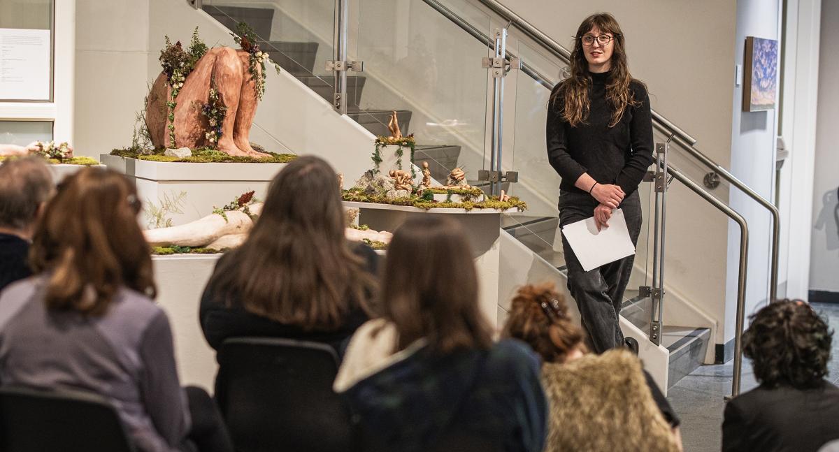 An audience is seated in front of a woman presenting alongside a series of sculptures
