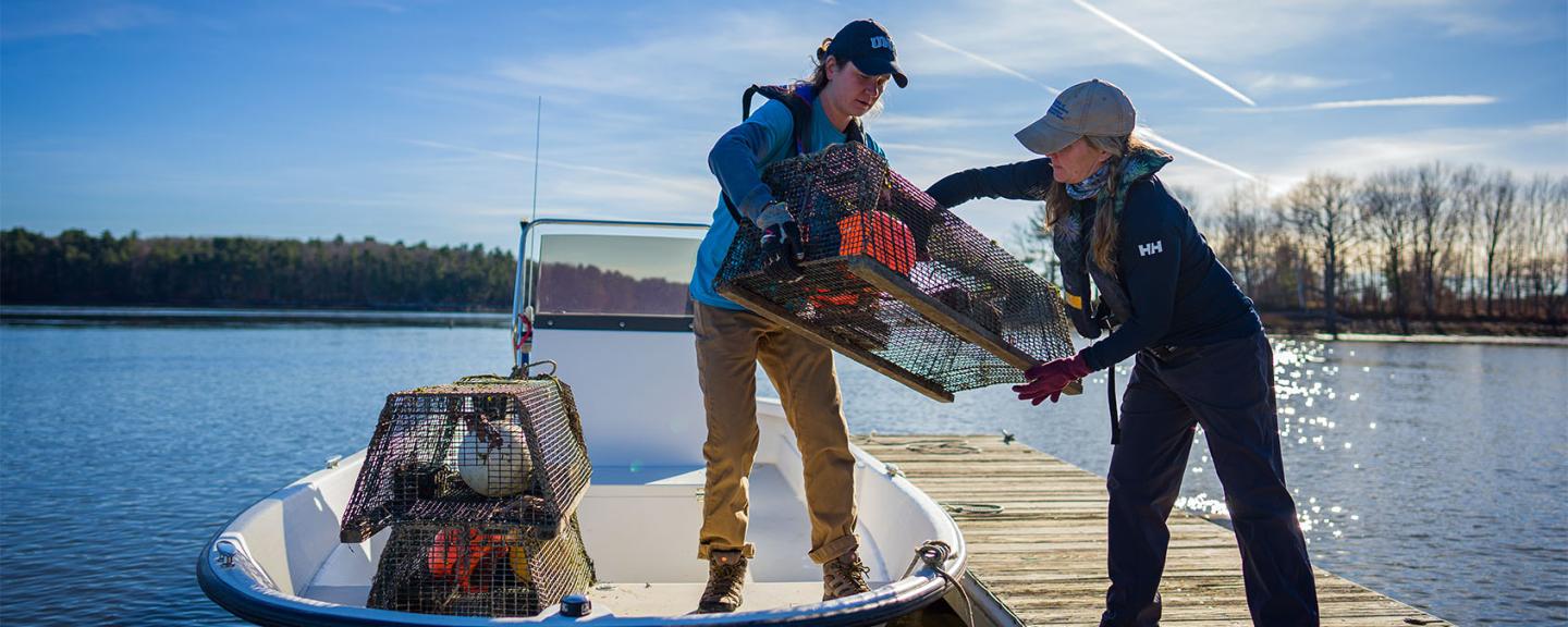 Two women stand on a dock, one on a boat (left) and the other on the dock (right). The woman on the boat hands the woman on the dock a trap used for shellfish. 