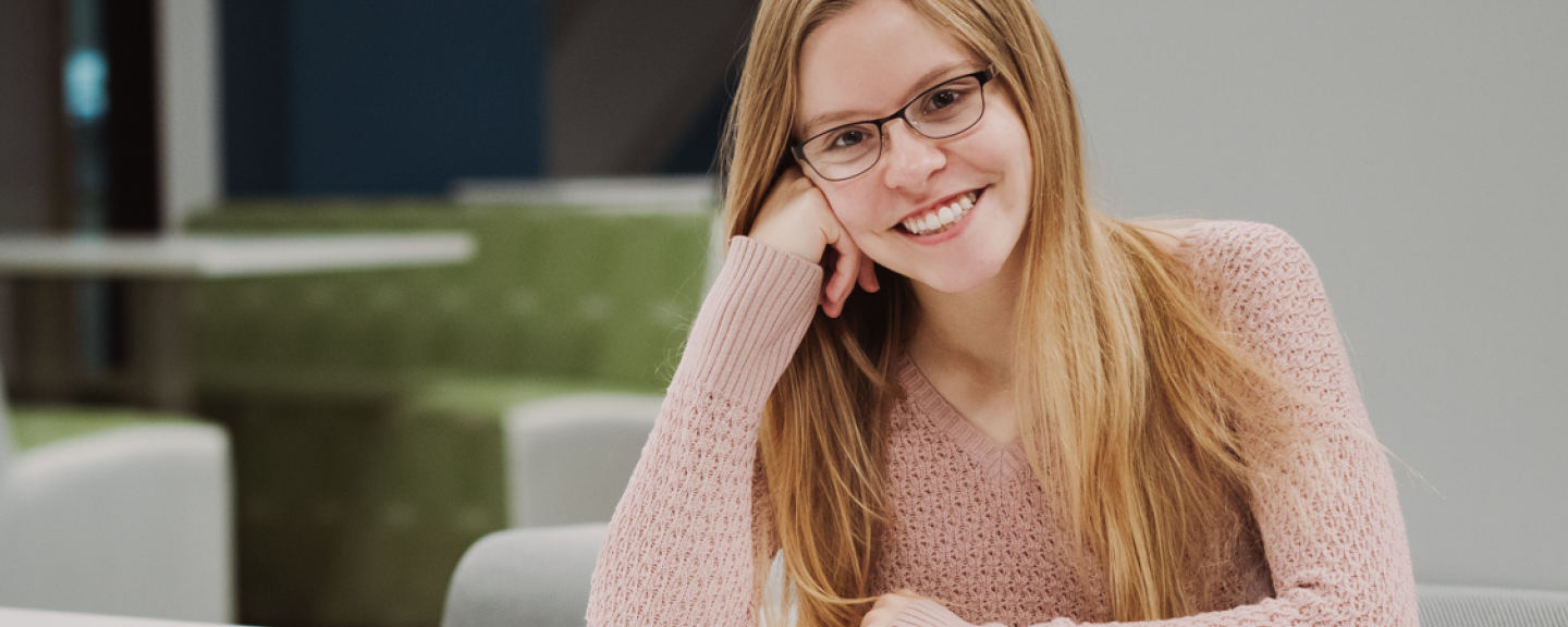 Paul College student smiles, leaning on a table. Booth seating is blurred in the background. She is wearing glasses, a pink sweater, and has strawberry blonde hair.