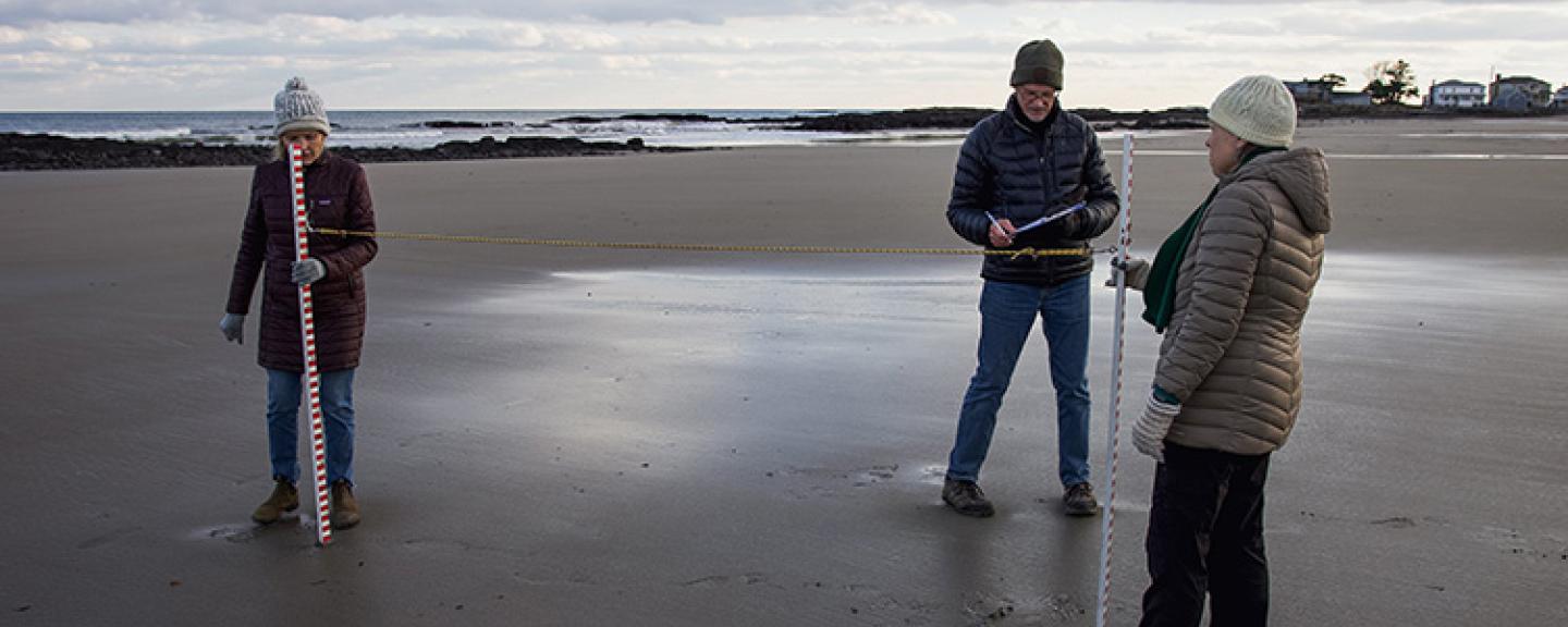 Two women holding yard sticks stand on wet sand in front of the ocean