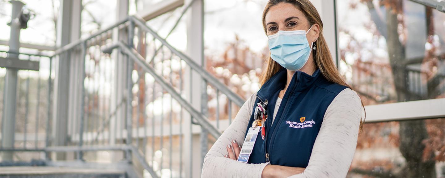 Nurse wearing a surgical mask standing in a glass-walled stairwell with a view of trees with their arms folded