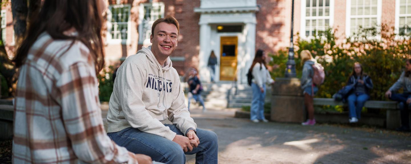 A male student in a white UNH sweatshirt talks to a female student in a flannel shirt with her back to the camera, as other students interact behind them