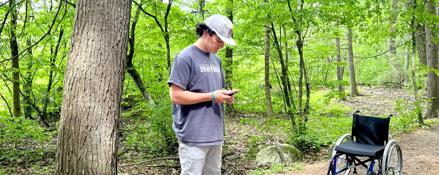 A man stands on a trail and uses his phone to record data while looking at levels laying on the ground. A wheelchair is in the background.