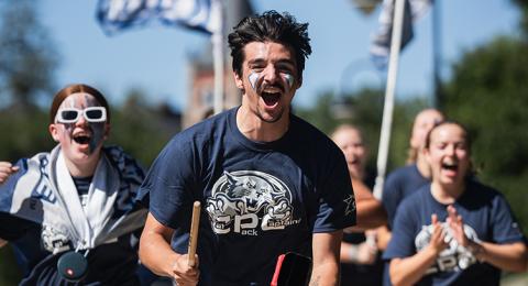 Male student with a cowbell and drumstick runs in front of a group of other cheering students