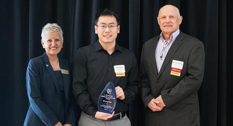 A man holding an award stands between a woman on the left and a man on the right