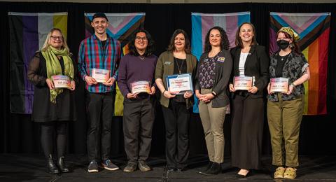 Group of seven award winners pose together on stage holding their awards
