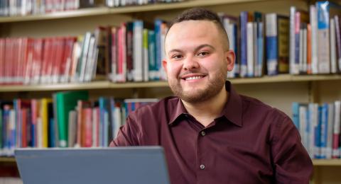 Photo of a man sitting in front of a laptop with a wall of bookshelves behind him. 