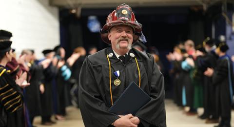 photo of a man wearing a fireman hat and graduation gown at a commencement ceremony