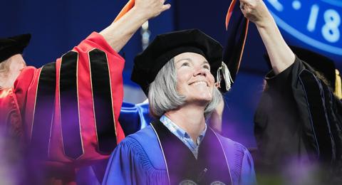 A woman looks up as an academic hood is lowered over her shoulders during a graduation ceremony