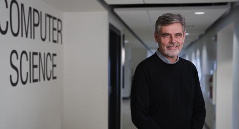 A man with graying hair and gray in his beard wearing a dark sweater stands in a hallway next to a wall labeled "Computer Science"