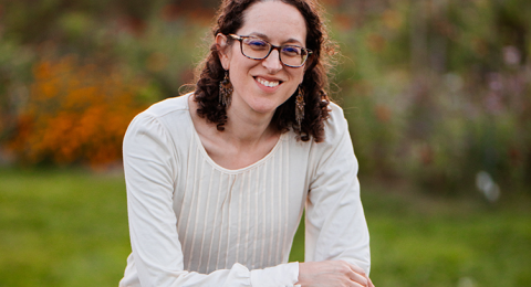 Portrait of a woman in glasses wearing a white sweater, sitting atop a picnic table in front of a wooded backdrop