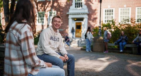A male student in a white UNH sweatshirt talks to a female student in a flannel shirt with her back to the camera, as other students interact behind them