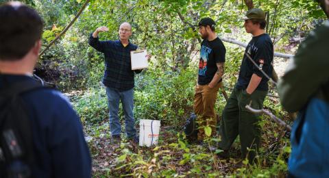 A man wearing a flannel shirt and glasses and holding a clipboard speaks to a group of students gathered around him in the woods