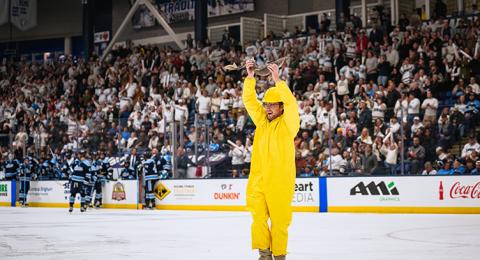 A man in a yellow fisherman outfit holds a fish over his head while standing on the ice in a hockey arena