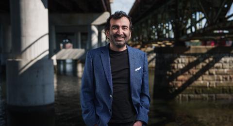 Man with dark hair, beard, and blue sportcoat stands under a highway bridge