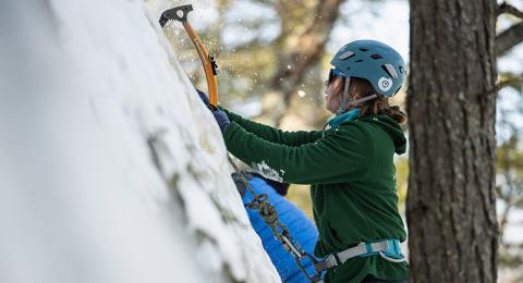 A woman in a helmet and green sweatshirt drives a climbing tool into an icy cliff face as she climbs up