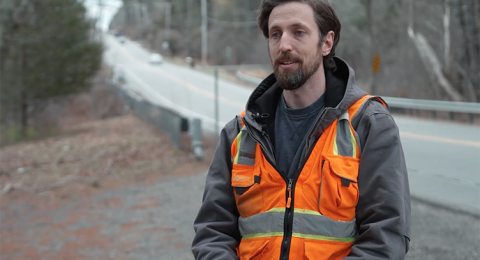 Screenshot from a video of a UNH researcher in an orange safety vest talking alongside a roadway