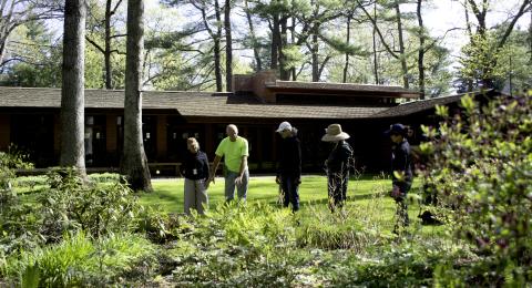 A group of people, dressed in gardening gear, stands near a perennial garden while a home is seen in the background.