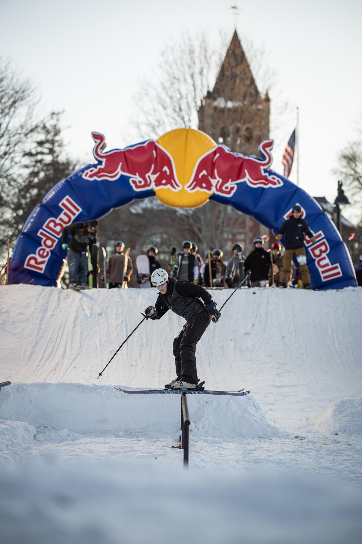 A crowd looks on beneath a Red Bull branded arch as a skier glides sideways along a rail