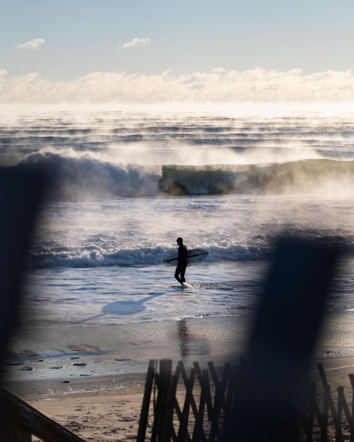 Steam rises from a series of waves as a surfer walks along the shoreline