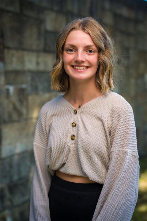 Master of Science in Nutrition student Kendra Perry. Kenda has white skin, blondish-brown hair that's cropped above her shoulders. She wears a greyish-white top and has black pants on.