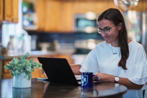 A woman with dark hair wearing glasses works at a computer on a circular table with a kitchen in the background