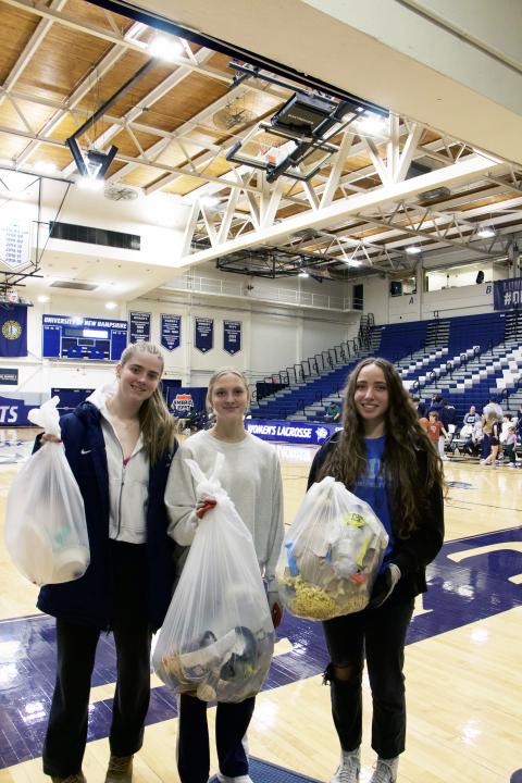 Three students holding trash bags with a basketball court and bleachers in the background 