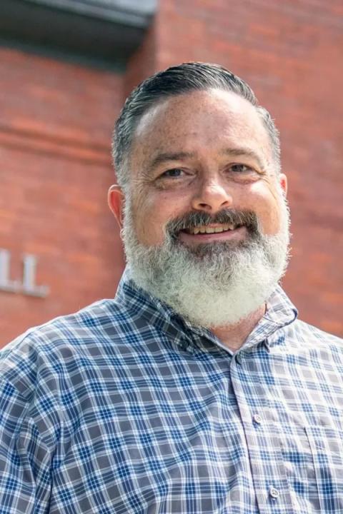 Portrait of Ken La Valley, with a white beard and wearing a blue and gray checkered shirt in front of a brick building