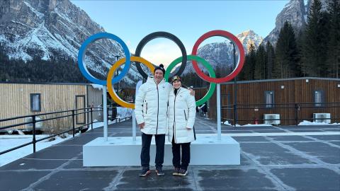 A man and woman in matching white puffer jackets stand in front of the Olympic rings and snow-capped mountains