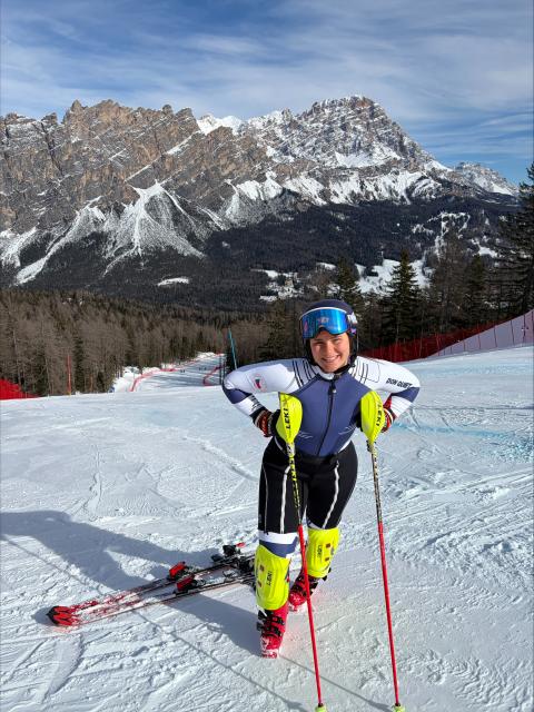A woman in a ski racing suit leans on her poles with her skis just behind her, while standing in front of snow-capped mountains