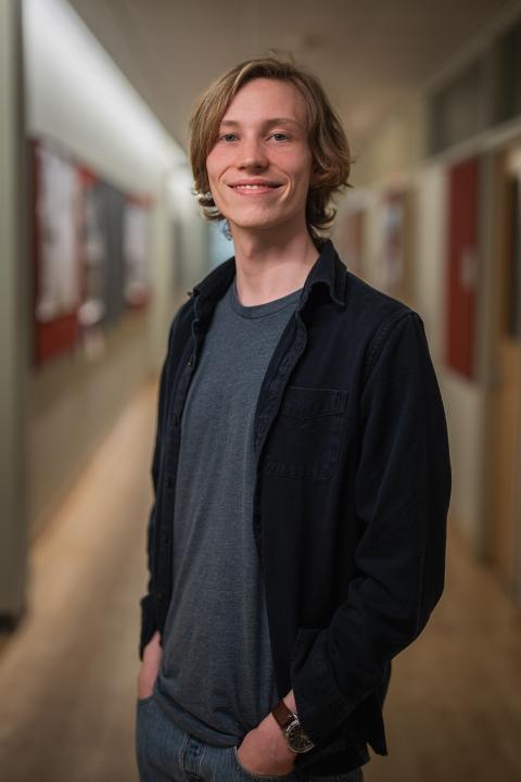 A male student in a black shirt over a gray T-shirt smiles while standing in a hallway