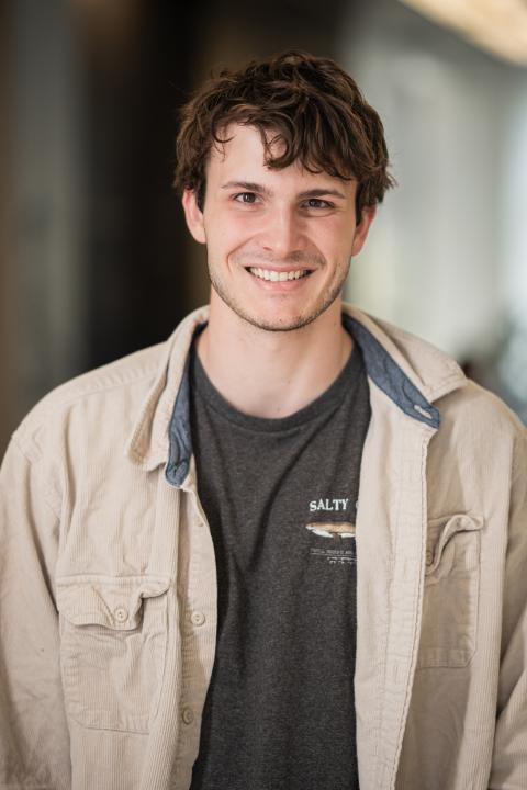 A male student in a light white jacket over a gray T-shirt smiles