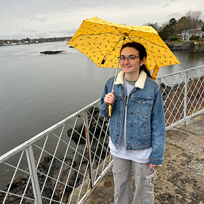UNH junior, Jess Calarese standing outside on a sidewalk by the water holding an umbrella