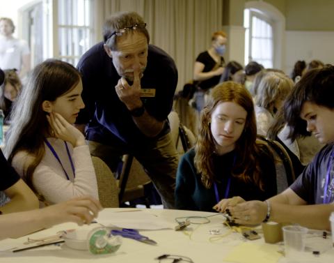 A group of teenagers work on an engineering puzzle while a man observes