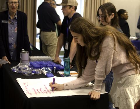 A young woman writes on a large sheet of paper draped over a table in a crowded room