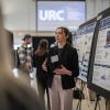 A female student stands in front of a poster explaining its contents to a few people standing in front of her